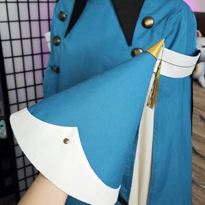 May include: A teal jacket with a black collar and gold-toned buttons, featuring a wide white cuff on the sleeve. The sleeve is accented with a pointed gold-colored cap and a thin gold tassel. The garment is displayed on a mannequin torso.