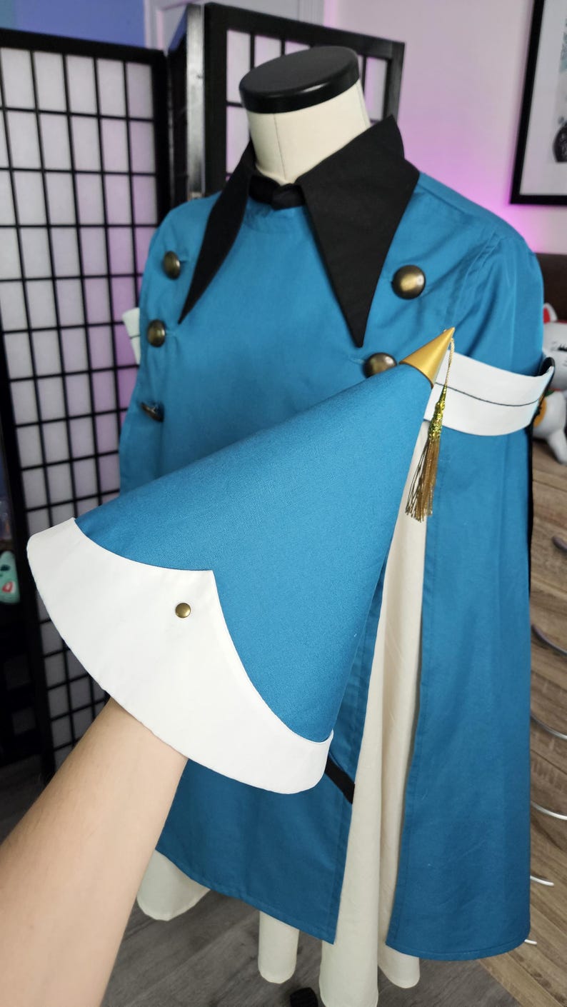 May include: A teal jacket with a black collar and gold-toned buttons, featuring a wide white cuff on the sleeve. The sleeve is accented with a pointed gold-colored cap and a thin gold tassel. The garment is displayed on a mannequin torso.