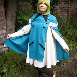 May include: A teal blue cape with a double-breasted front, black lapels, and white sleeve cuffs worn over a white dress. The outfit includes a matching teal blue pointed hat with a gold tip, dark brown leggings, and brown flat shoes.