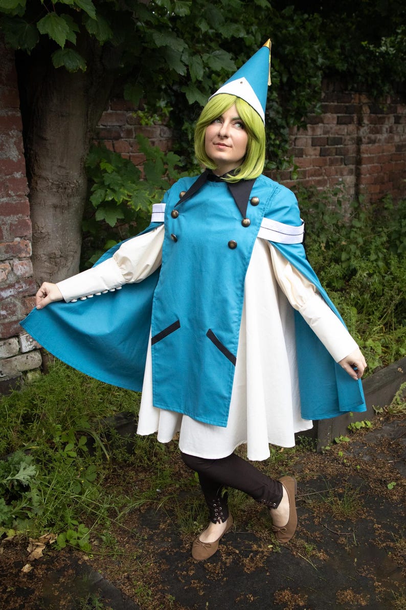 May include: A teal blue cape with a double-breasted front, black lapels, and white sleeve cuffs worn over a white dress. The outfit includes a matching teal blue pointed hat with a gold tip, dark brown leggings, and brown flat shoes.