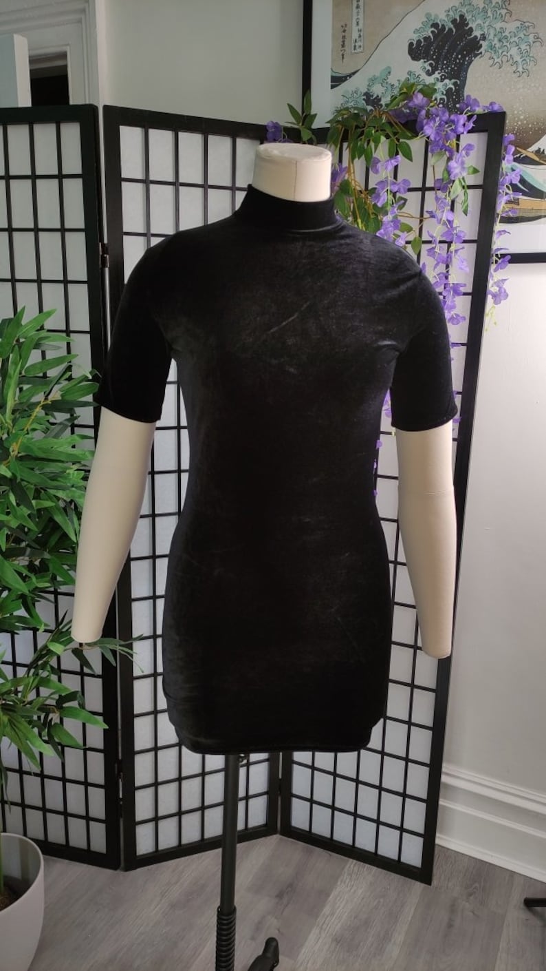 Puede incluir: Un minivestido negro de terciopelo con mangas cortas y cuello alto. El vestido se exhibe en un maniqu&iacute;. El vestido es de color negro liso y tiene un aspecto suave y texturizado. El vestido es de estilo ajustado.
