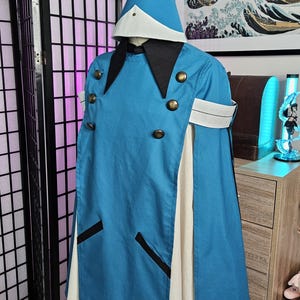 May include: A blue hooded cape with a white underlayer, featuring a pointed hat with a gold tassel, black collar, and six brass-toned buttons. The garment includes white arm bands and two black-trimmed pockets on the front of the blue overlay.
