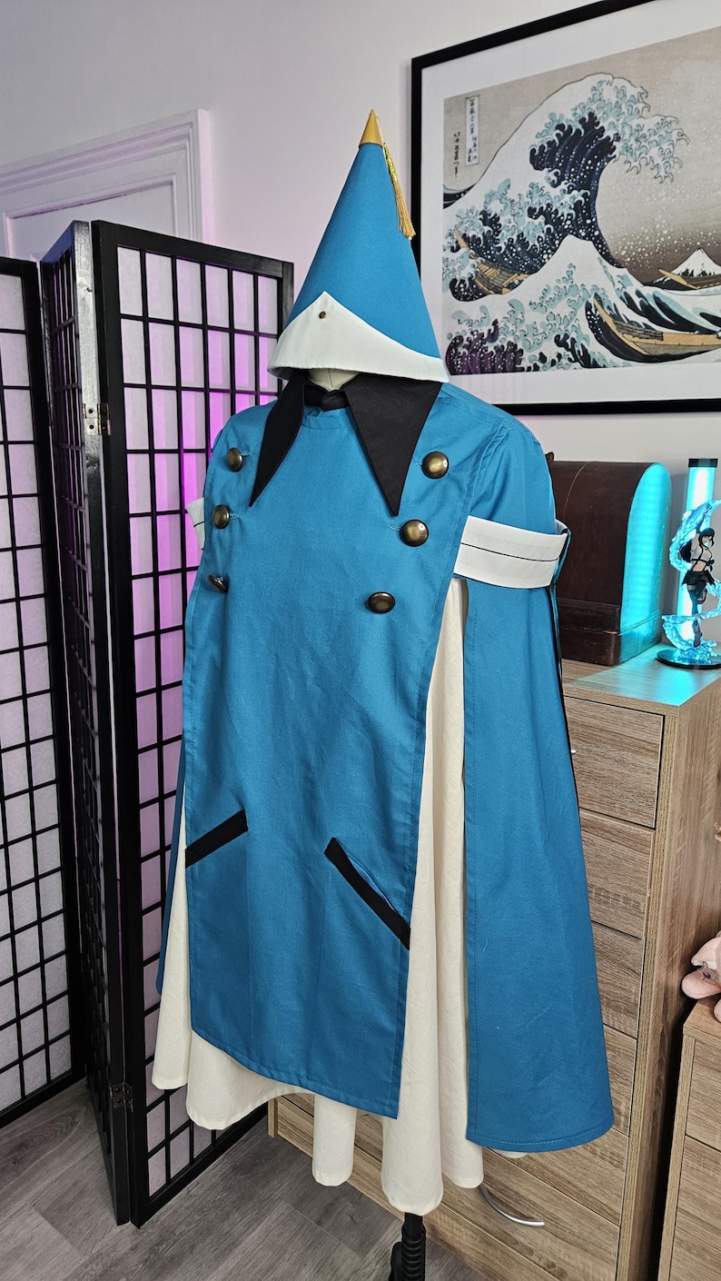 May include: A blue hooded cape with a white underlayer, featuring a pointed hat with a gold tassel, black collar, and six brass-toned buttons. The garment includes white arm bands and two black-trimmed pockets on the front of the blue overlay.