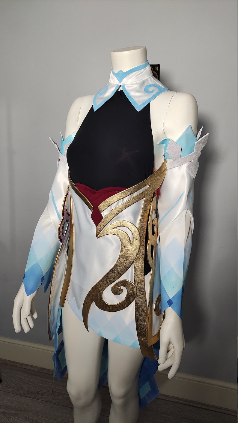 May include: A white and blue cosplay costume with gold accents. The costume features a black top, a white skirt with a blue and white pattern, and gold trim. The costume is worn on a mannequin.