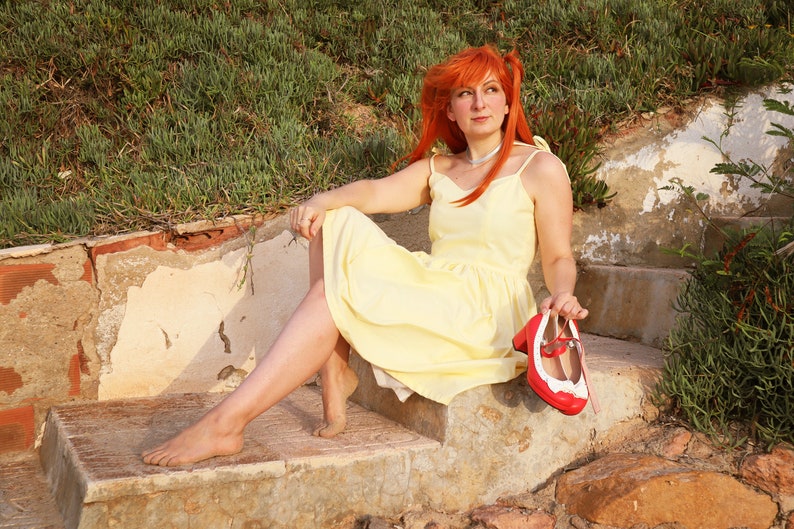 K&ouml;nnte beinhalten: Eine Frau mit leuchtend roten Haaren sitzt auf einer Steintreppe und tr&auml;gt ein gelbes Sommerkleid und rote Schuhe. Sie h&auml;lt einen der Schuhe in der Hand.