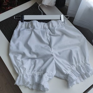 May include: A pair of white bloomers featuring an elasticized waistband and gathered leg openings finished with a delicate white eyelet lace trim. The garment is displayed on a white plastic hanger, showcasing a loose, voluminous silhouette.
