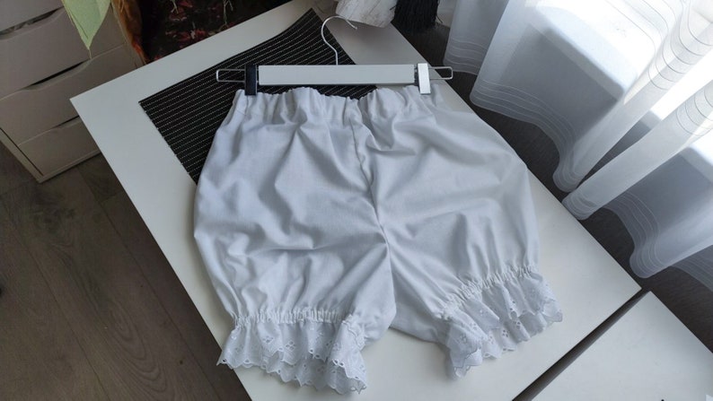 May include: A pair of white bloomers featuring an elasticized waistband and gathered leg openings finished with a delicate white eyelet lace trim. The garment is displayed on a white plastic hanger, showcasing a loose, voluminous silhouette.