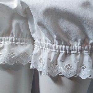 May include: White bloomers featuring a gathered elastic waistband at the leg openings and a decorative eyelet lace trim with a scalloped edge. The fabric is a solid white cotton material with a soft, gathered texture above the hemline.