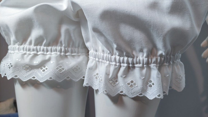 May include: White bloomers featuring a gathered elastic waistband at the leg openings and a decorative eyelet lace trim with a scalloped edge. The fabric is a solid white cotton material with a soft, gathered texture above the hemline.