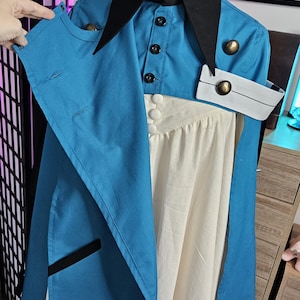 May include: A blue and cream-colored dress featuring a black pointed collar, a button-up bodice, and a blue overcoat layer with black trim. The garment includes metallic gold buttons, a white cuff detail, and a pleated cream skirt section.