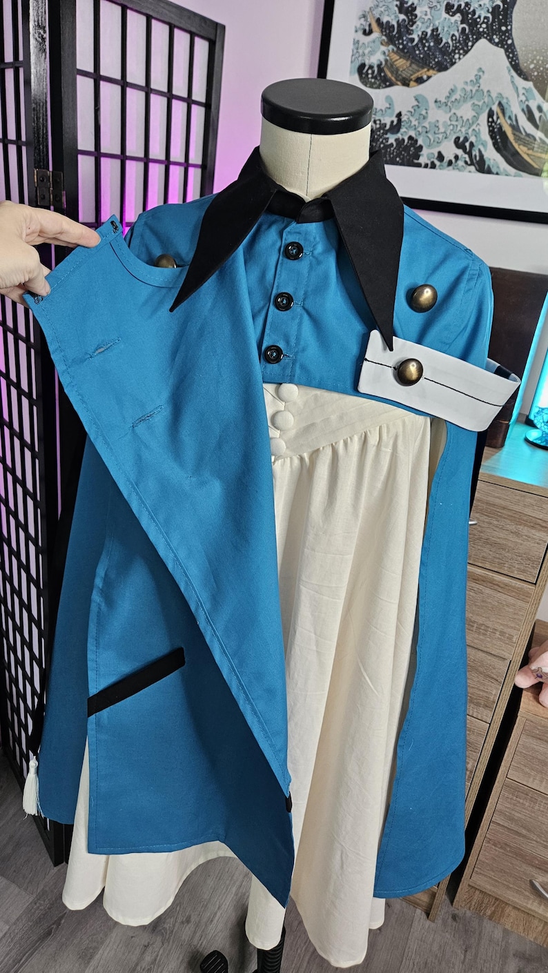 May include: A blue and cream-colored dress featuring a black pointed collar, a button-up bodice, and a blue overcoat layer with black trim. The garment includes metallic gold buttons, a white cuff detail, and a pleated cream skirt section.