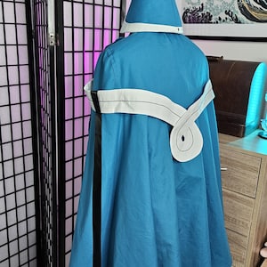 May include: A blue hooded cloak featuring a pointed hat with a gold-colored tip and tassel. The cloak includes a white sash tied around the waist with a long black ribbon hanging down the side, worn over a white garment with a flared hemline.
