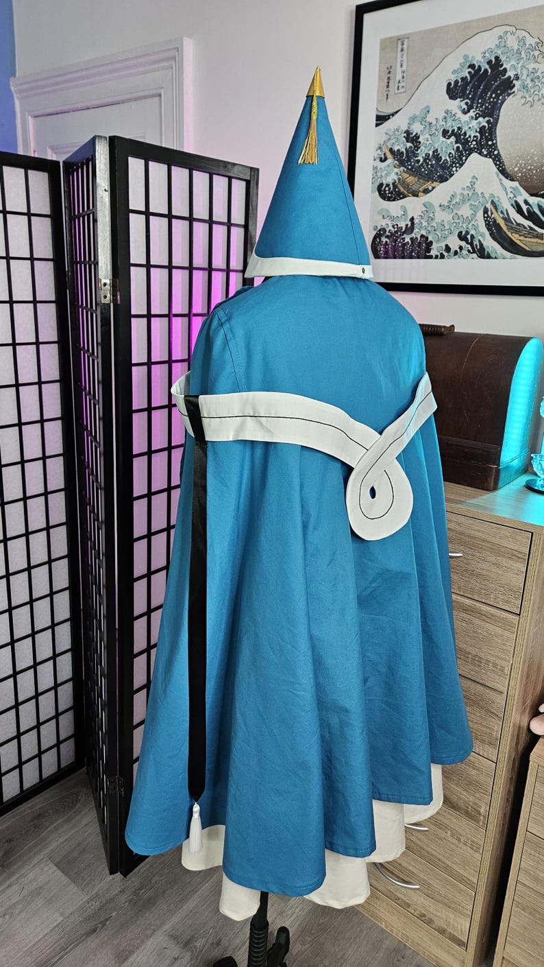 May include: A blue hooded cloak featuring a pointed hat with a gold-colored tip and tassel. The cloak includes a white sash tied around the waist with a long black ribbon hanging down the side, worn over a white garment with a flared hemline.