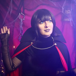 May include: A black cape with a high collar and a vibrant red lining, secured at the neck with a gold-toned chain. The garment is worn over a black long-sleeved top with sheer mesh sleeves, featuring a dark aesthetic with a dramatic, structured silhouette.