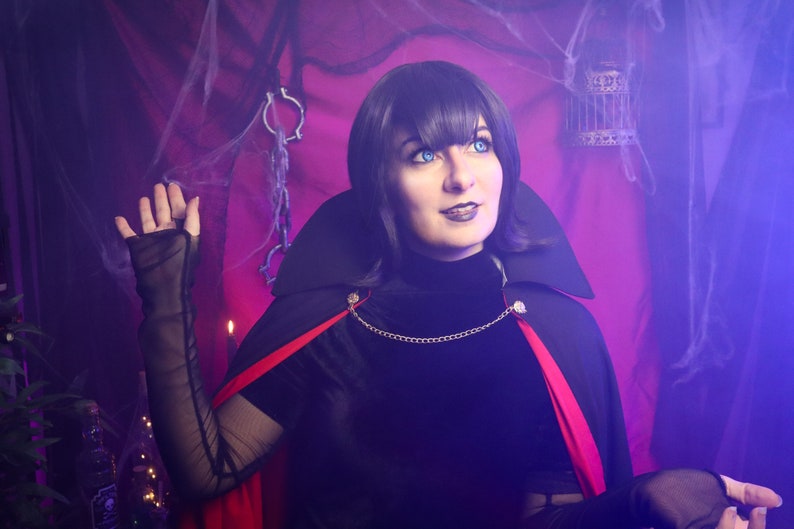 May include: A black cape with a high collar and a vibrant red lining, secured at the neck with a gold-toned chain. The garment is worn over a black long-sleeved top with sheer mesh sleeves, featuring a dark aesthetic with a dramatic, structured silhouette.
