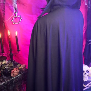 May include: A solid black cape featuring a high, structured collar that stands upright around the neck. The garment is floor-length and drapes down the back, constructed from a smooth, opaque fabric that provides full coverage from the shoulders down.