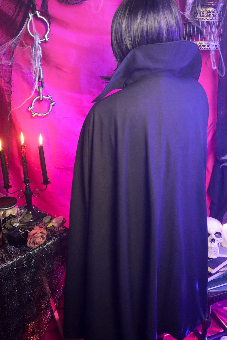 May include: A solid black cape featuring a high, structured collar that stands upright around the neck. The garment is floor-length and drapes down the back, constructed from a smooth, opaque fabric that provides full coverage from the shoulders down.