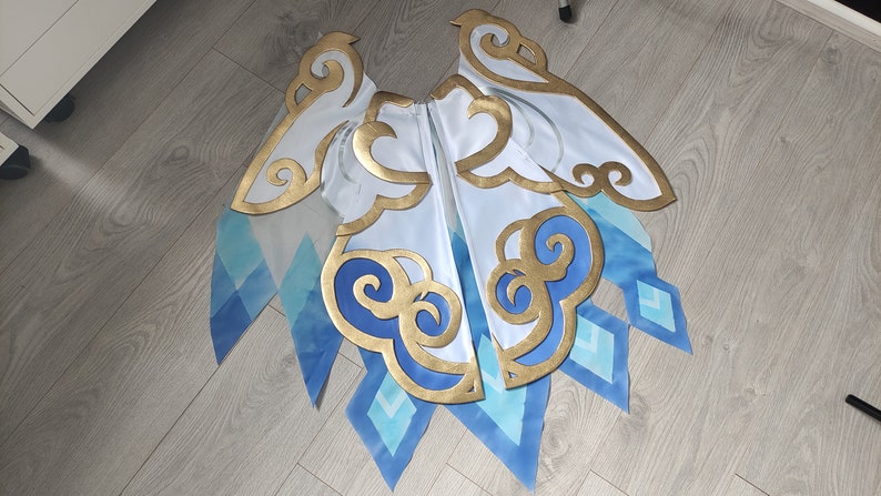 May include: A white fabric costume piece with blue and gold accents. The fabric has a cloud and diamond pattern. The piece is designed to look like wings.