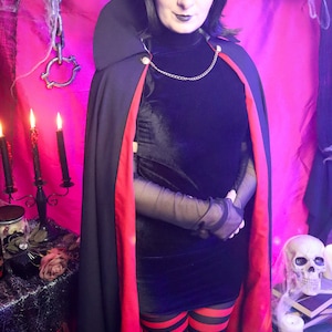 May include: A black velvet dress with a high neck, sheer black sleeves, and a black cape featuring a red lining and a gold chain closure. The outfit includes red and black horizontal striped leggings, paired with a short black bob hairstyle.