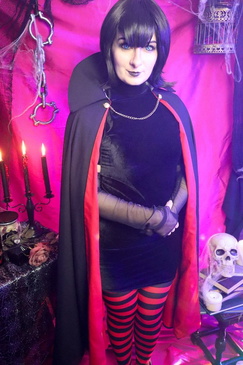 May include: A black velvet dress with a high neck, sheer black sleeves, and a black cape featuring a red lining and a gold chain closure. The outfit includes red and black horizontal striped leggings, paired with a short black bob hairstyle.