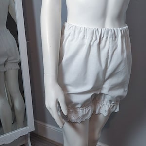 May include: White bloomers featuring an elasticized waistband and leg openings finished with delicate eyelet lace trim. These shorts are crafted from a solid white fabric and provide a loose, comfortable fit with gathered detailing at the hem.