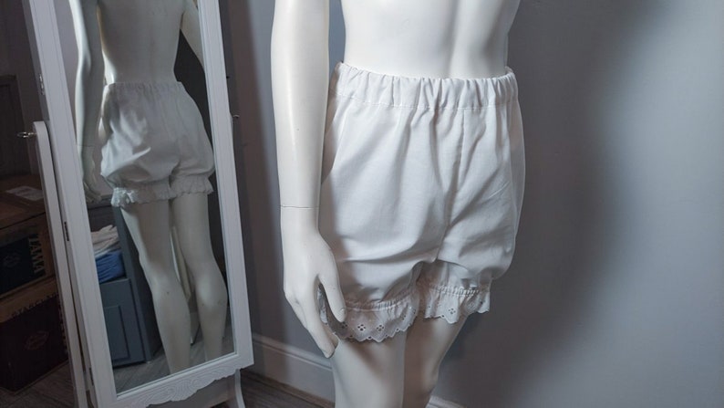 May include: White bloomers featuring an elasticized waistband and leg openings finished with delicate eyelet lace trim. These shorts are crafted from a solid white fabric and provide a loose, comfortable fit with gathered detailing at the hem.