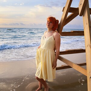 K&ouml;nnte beinhalten: Eine Frau mit roten Haaren tr&auml;gt ein gelbes Sommerkleid und steht an einem Sandstrand mit dem Meer im Hintergrund. Sie lehnt sich an einen h&ouml;lzernen Rettungsturm.