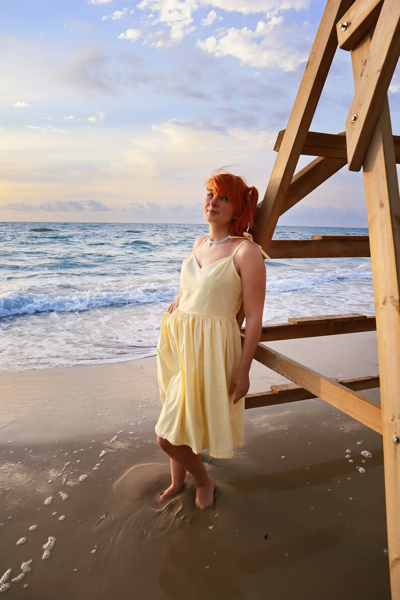 K&ouml;nnte beinhalten: Eine Frau mit roten Haaren tr&auml;gt ein gelbes Sommerkleid und steht an einem Sandstrand mit dem Meer im Hintergrund. Sie lehnt sich an einen h&ouml;lzernen Rettungsturm.