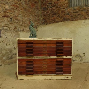 May include: A wooden plan chest with two wide drawers, each featuring five horizontal wooden pulls and brass label holders. The cabinet has a distressed cream-coloured frame and a dark wood finish on the drawer fronts. A small green hare figurine sits on top.