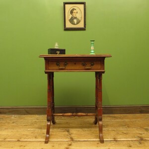 May include: An antique wooden side table with two drawers and decorative legs. A framed portrait hangs above the table, and a green candle holder and a box are on top. The table is against a green wall.
