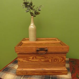 May include: A wooden storage chest with a metal handle and a light-coloured bottle holding greenery on top. The chest is rectangular and made of light brown wood. The bottle is beige.