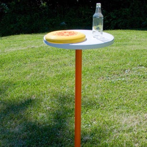 May include: A small, round white table with an orange pole and a yellow frisbee on top. A clear glass bottle sits next to the frisbee. The table is outdoors on a grassy lawn.