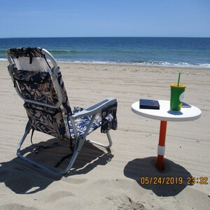Puede incluir: Una escena de playa con una silla de playa plegable con un estampado floral azul y blanco, una pequeña mesa blanca y un vaso verde con una pajita. La mesa tiene un poste naranja y blanco. El océano está al fondo.