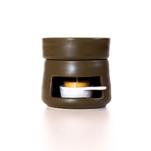 May include: A dark olive green ceramic wax warmer with a square opening and a small white dish inside. A lit yellow tea light candle sits in the dish. The warmer has a two-piece design.