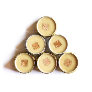 May include: Six small round containers with lids, arranged in a triangle shape. Each container is filled with a light beige substance and has a small square of gold leaf on top.