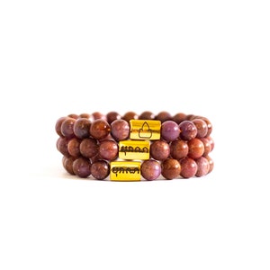 May include: Three stacked bracelets made of red and brown gemstone beads with gold accents. Each gold accent has a different inscription in a foreign language.