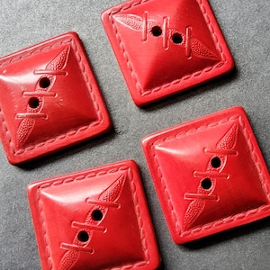 May include: Four red plastic buttons with a stitched design. Each button has a square shape with rounded corners and a raised stitched design that resembles a seam.