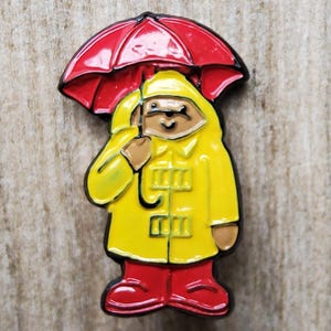 Könnte beinhalten: Ein gelber Regenmantel mit einem roten Regenschirm und roten Stiefeln. Der Regenmantel hat eine Kapuze und Knöpfe. Der Regenschirm ist geöffnet und hat einen roten Griff.