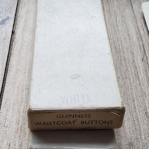 May include: A rectangular white box labeled "WHITE" and "GUINNESS WAISTCOAT BUTTONS" in black text. The box is sitting on top of a smaller, similar box. The boxes are on a wooden surface.