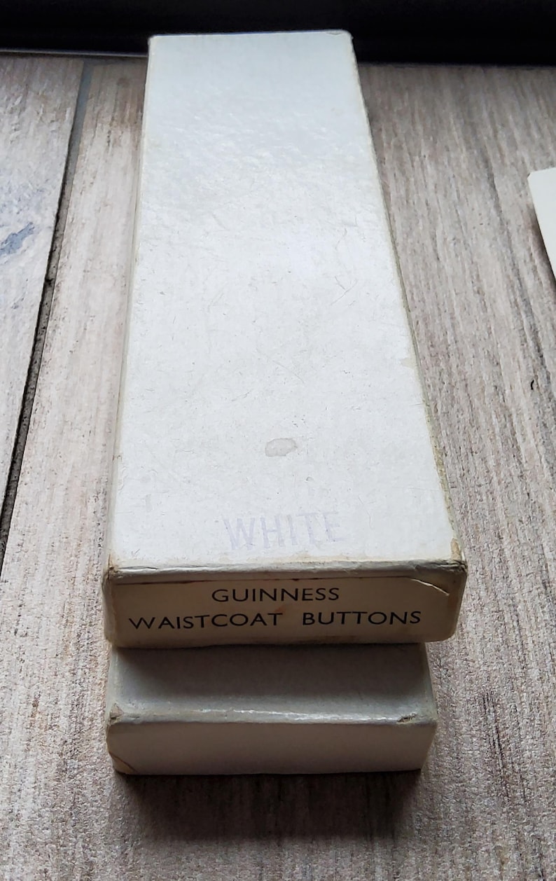 May include: A rectangular white box labeled "WHITE" and "GUINNESS WAISTCOAT BUTTONS" in black text. The box is sitting on top of a smaller, similar box. The boxes are on a wooden surface.