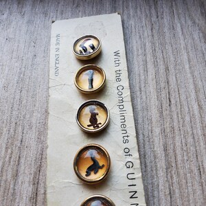 May include: A set of five vintage buttons with gold-toned rims and domed, clear centers. Each button features a different black silhouette design on an amber background. The card reads "Made in England" and "With the Compliments of GUINN".