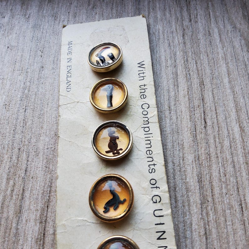 May include: A set of five vintage buttons with gold-toned rims and domed, clear centers. Each button features a different black silhouette design on an amber background. The card reads "Made in England" and "With the Compliments of GUINN".
