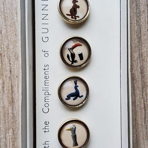 May include: Set of six vintage Guinness-themed buttons with gold-tone rims. Each button features a different whimsical illustration, including a turtle with a pint, a kangaroo, and a toucan. The text "With the Compliments of Guinness" is visible.