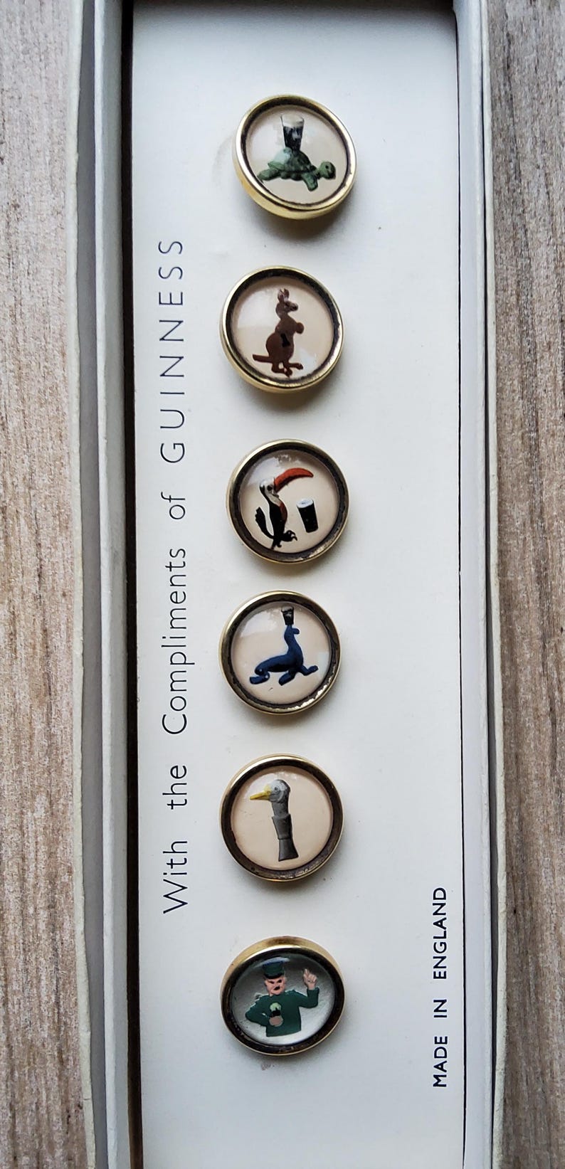 May include: Set of six vintage Guinness-themed buttons with gold-tone rims. Each button features a different whimsical illustration, including a turtle with a pint, a kangaroo, and a toucan. The text "With the Compliments of Guinness" is visible.