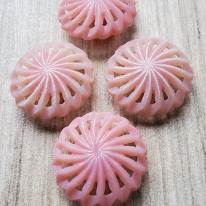 Könnte beinhalten: Fünf rosa, blütenförmige Perlen mit einem Spiraldesign. Die Perlen sind hellrosa, durchscheinend und haben ein detailliertes, geschnitztes Aussehen. Die Perlen sind auf einer hellen Holzoberfläche angeordnet.
