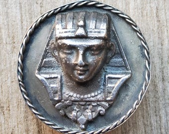 May include: Silver-toned metal belt buckle featuring a detailed embossed design of an Egyptian pharaoh's head wearing a crown.