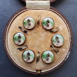Può includere: Un set di sei gemelli rotondi color oro con un'illustrazione dipinta a mano di una persona in bicicletta su un sentiero. Ogni gemello ha uno sfondo verde e bianco ed è esposto in una custodia circolare foderata in velluto marrone.