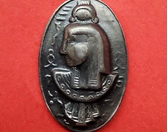 May include: A silver oval-shaped pendant featuring a raised image of a woman's head wearing a crown and headdress. The pendant is intricately detailed and surrounded by a border of text.