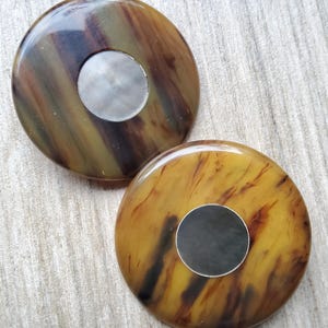 May include: Two round, decorative buttons with a marbled pattern in shades of brown, yellow, and black. Each button has a silver-colored, circular inset in the center.