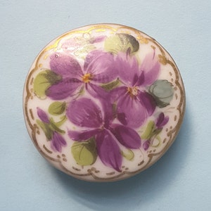 May include: A round, white porcelain button with a gold trim and a hand-painted design of purple violets and green leaves.